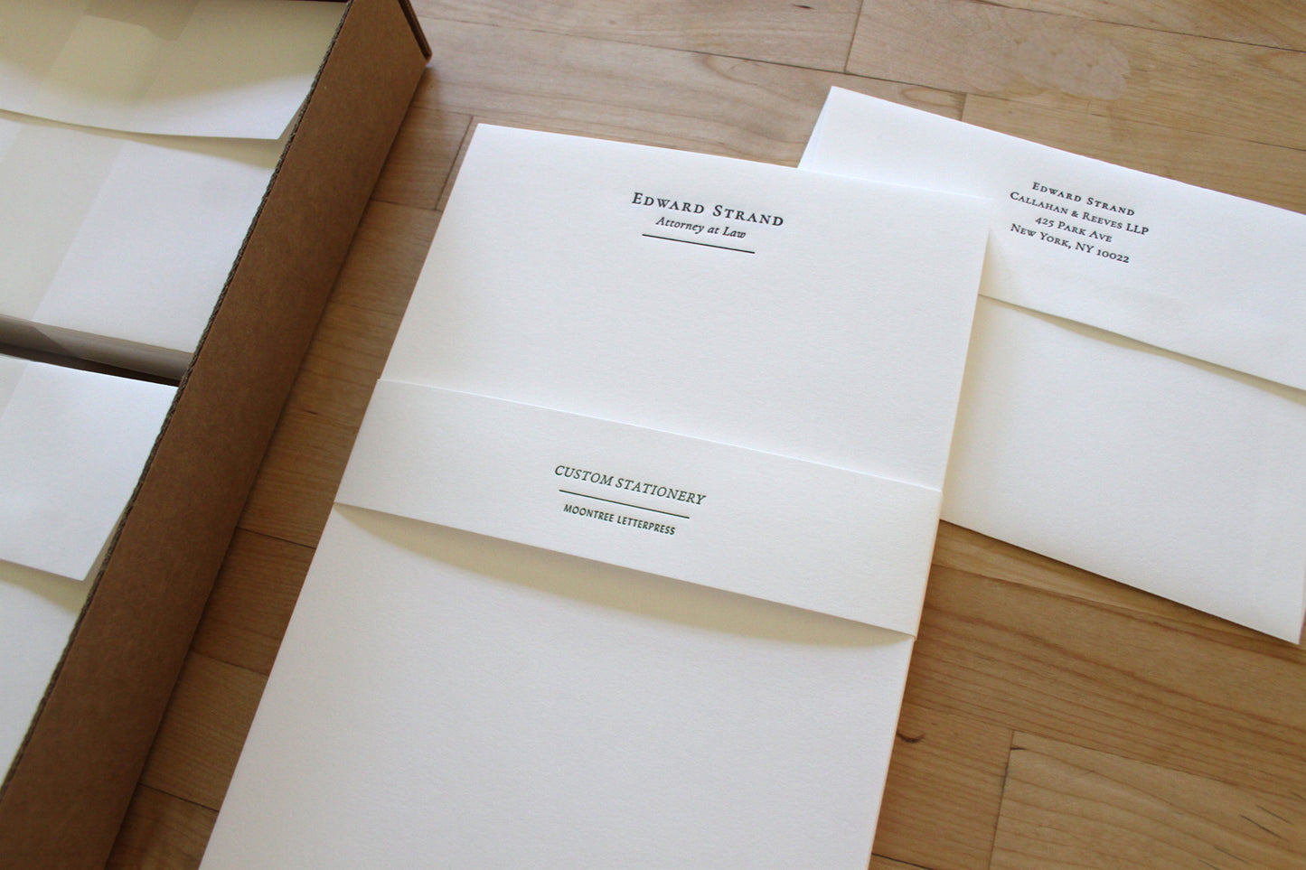 Custom letterpress stationery envelopes and cards with printed text laid out on a light wood floor, alongside a brown cardboard box.